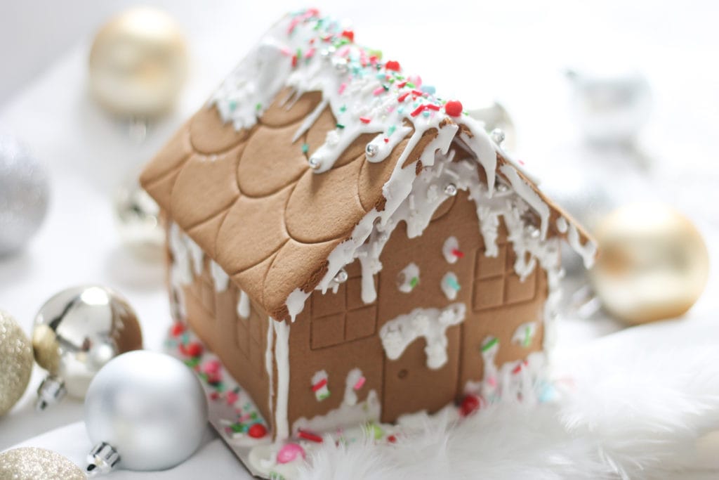 Finished Gingerbread House with icing and sprinkles on fluffy white tree skirt surrounded by ornaments