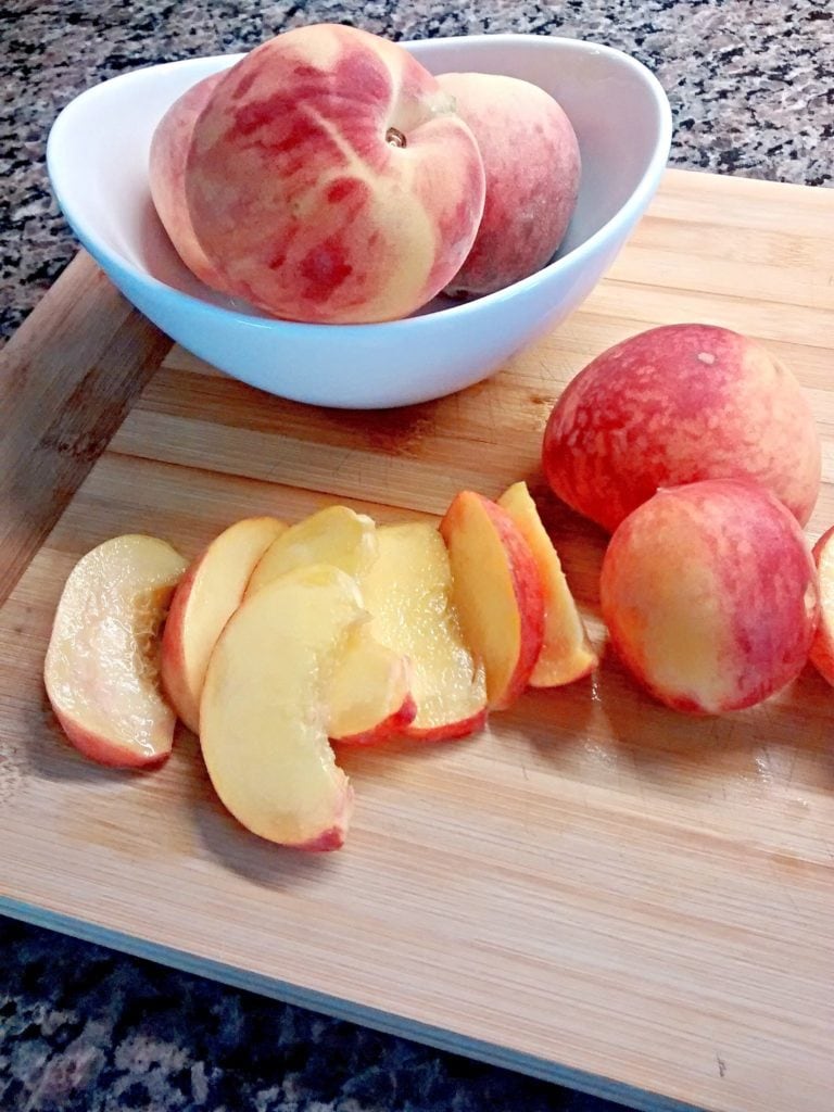 Fresh peaches sliced on cutting board with white ceramic bowl full of whole peaches
