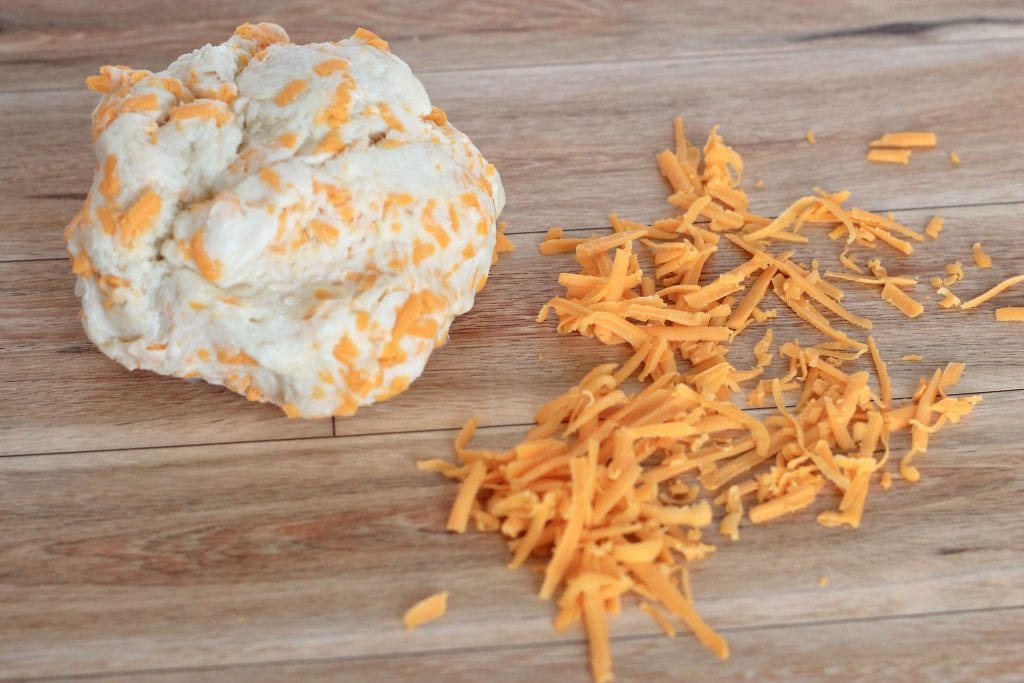 Shredded cheddar cheese and biscuit dough ball on wooden cutting board.