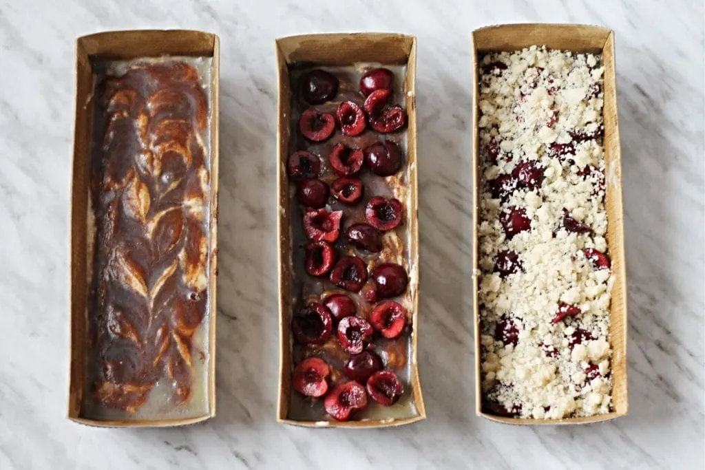 Three stages of Cherry Coffee Cake in individual baking trays. First has cinnamon swirl, second has sliced cherries and the final one is the streusel topping. 