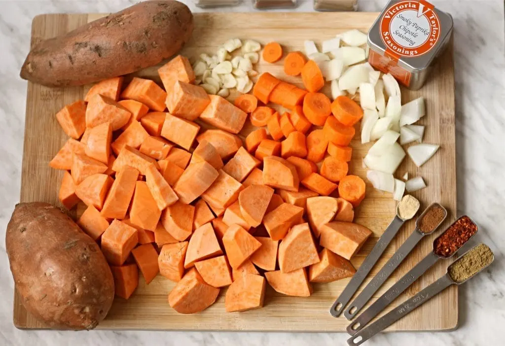 Wooden cutting board with diced sweet potatoes, carrots, onions and other ingredients to make sweet potato soup.