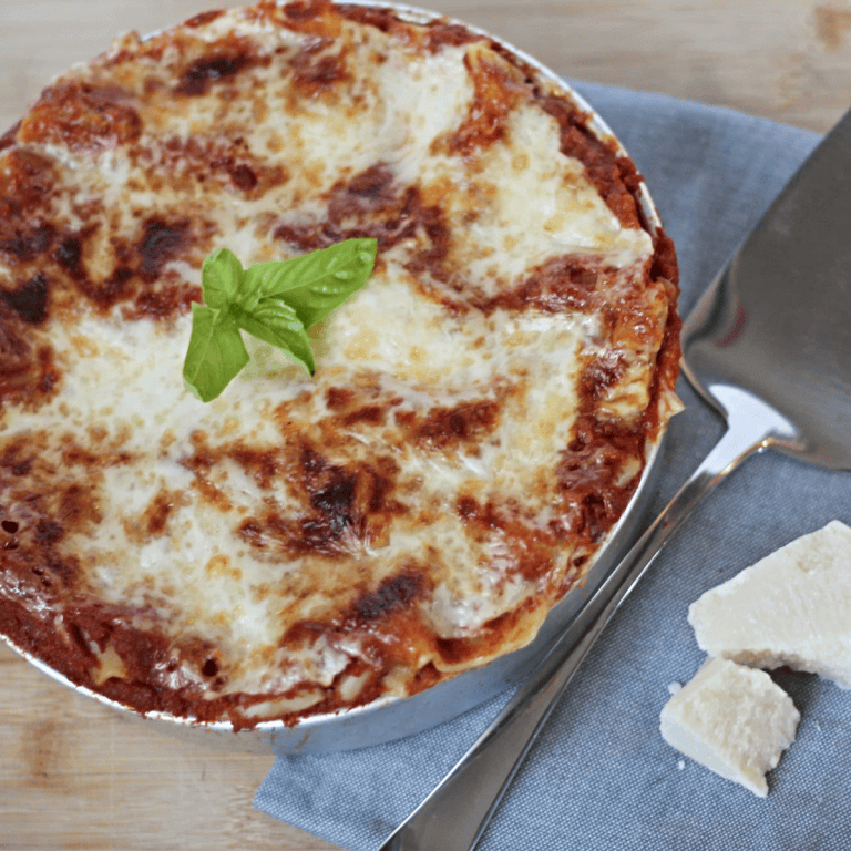 Round dish with lasagna, topped with fresh basil and a serving spoon and chunks of Parmesan cheese next to it.