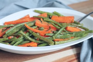 White serving dish filled with green beans and carrots on wooden cutting board