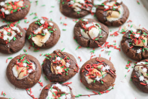 Fudgy Hot Chocolate Cookies on baking sheet topped with festive sprinkles, marshmallows and chocolate drizzle