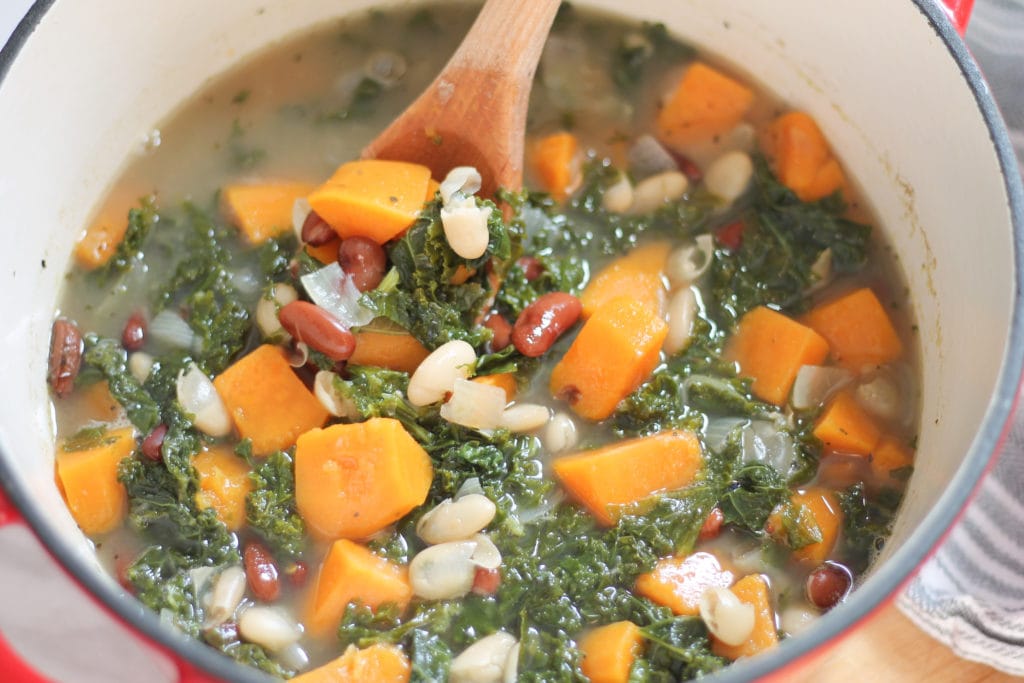Hearty Kale Soup with Sweet Potatoes and beans in red dutch oven with wooden spoon 
