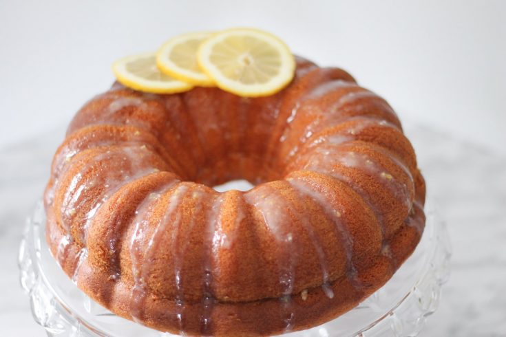 Finished lemon cake on crystal cake stand with freshly sliced lemons on top.