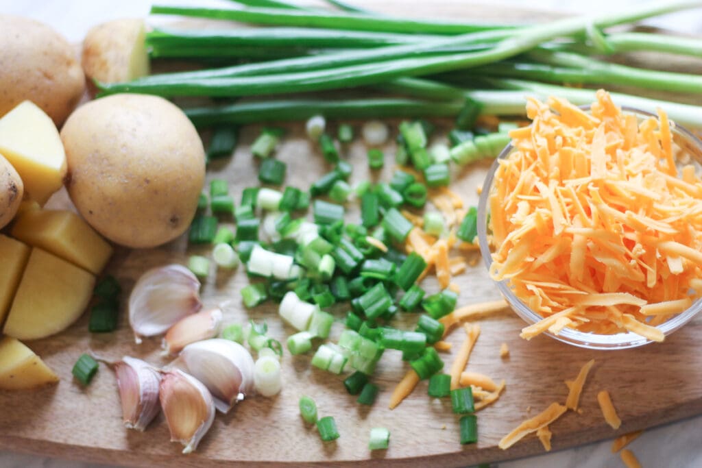 Wooden cutting board with potatoes, green onions, garlic, cheddar cheese and other ingredients needed to make the dish.