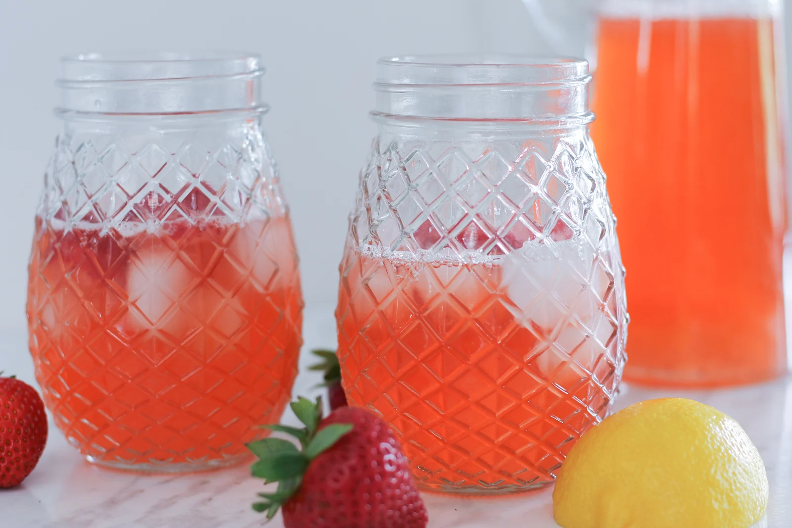 Two glass of fresh strawberry lemonade on countertop with pitcher and fresh strawberries and lemons beside them.