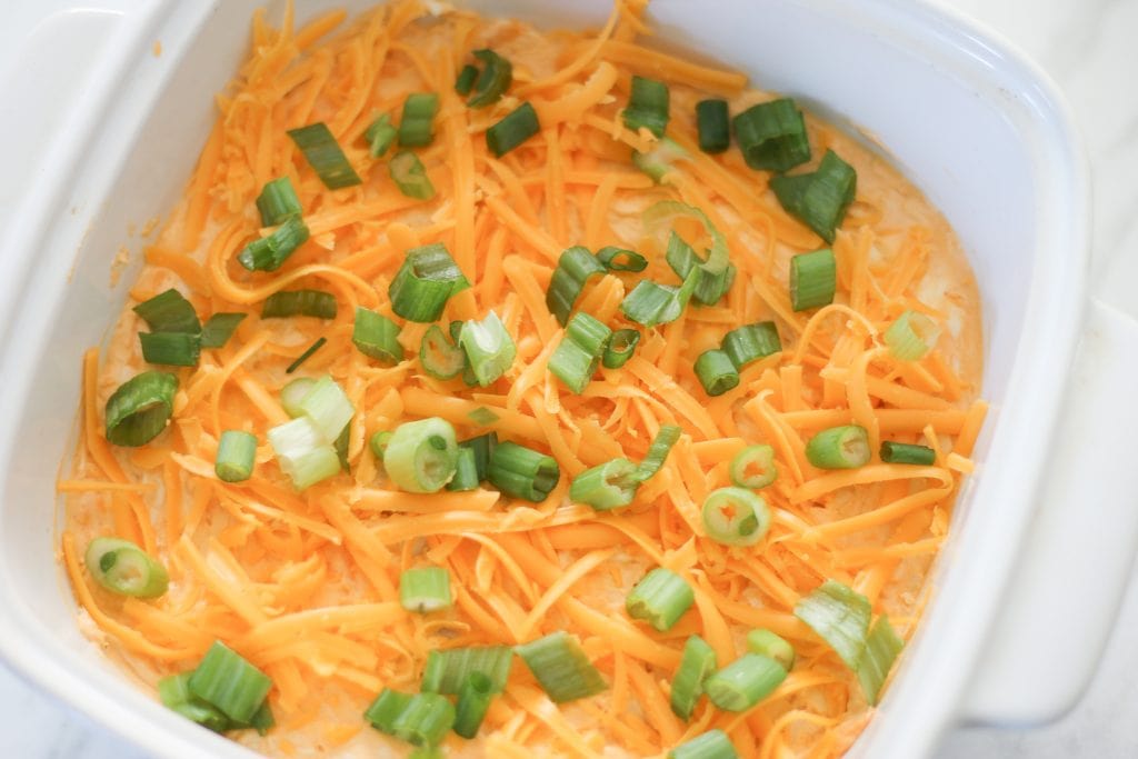 Assembled dip in white ceramic baking dish, topped with shredded cheese and sliced green onions.