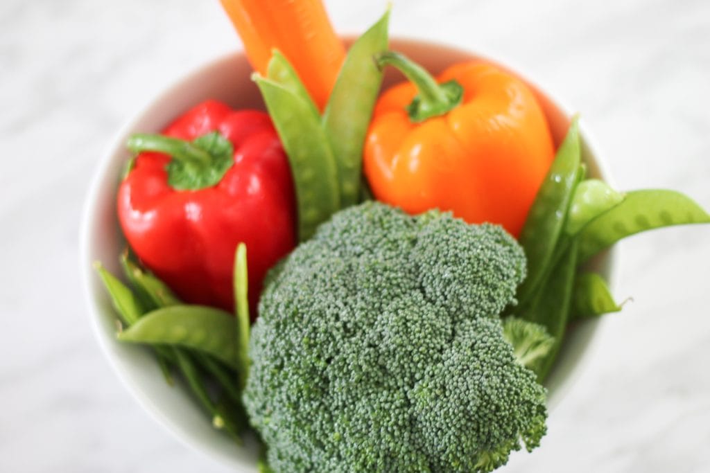 White bowl on counter filled with fresh vegetables like broccoli, carrots, peppers and peas.