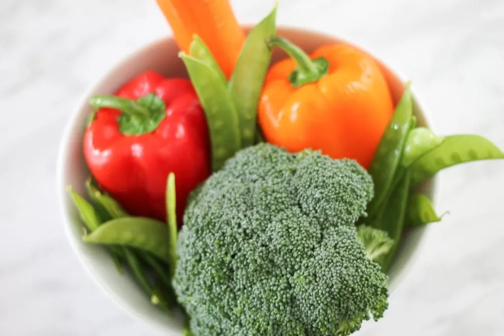 White bowl on counter filled with fresh vegetables like broccoli, carrots, peppers and peas.